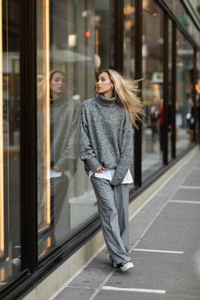 Повний зріст молодої і стилізованої жінки в сірому одязі, що стоїть біля віконної виставки в Нью-Йорку.  - Фото, зображення