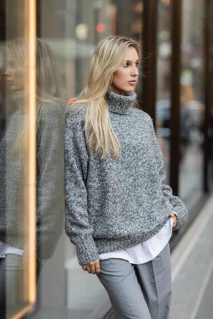 блондинка в зимнем наряде стоит с рукой в кармане возле витрины в Нью-Йорке  - Фото, изображение