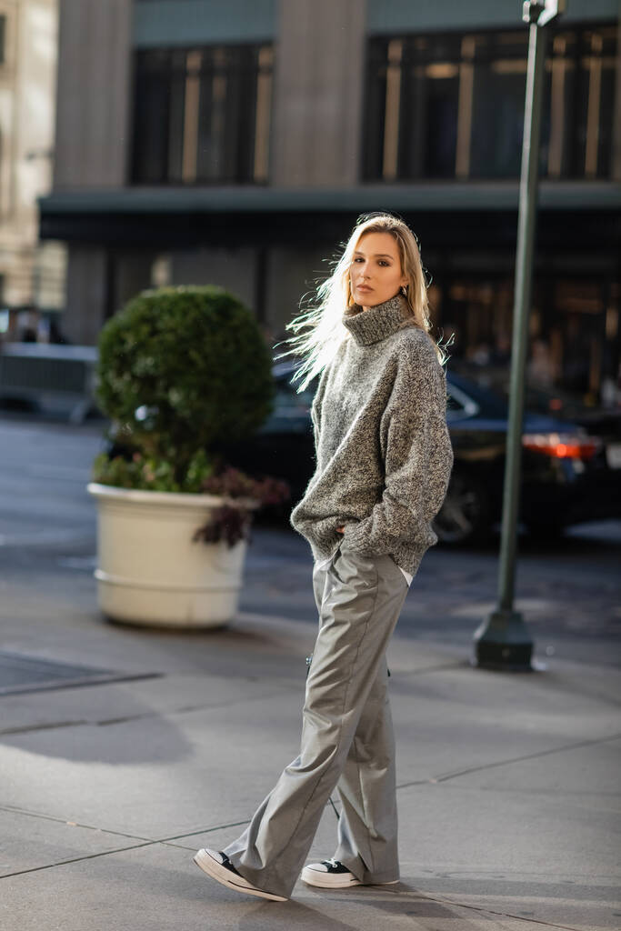 longitud completa de mujer joven con estilo con cabello rubio mirando a la cámara mientras está de pie con la mano en el bolsillo en la ciudad de Nueva York  - Foto, Imagen
