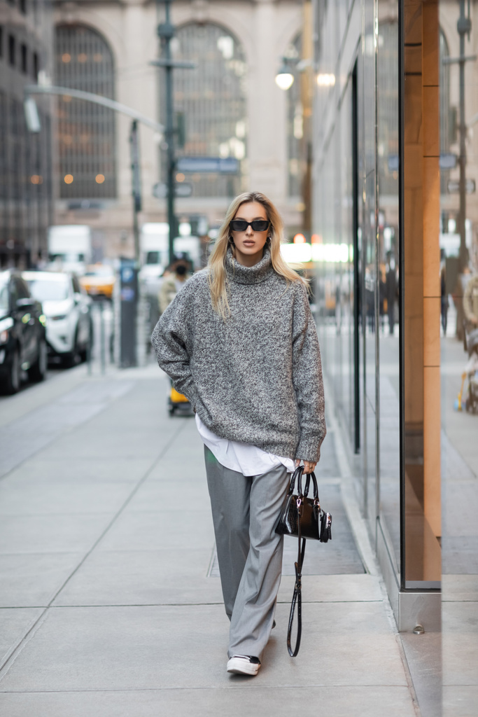 volledige lengte van blonde vrouw in de winter trui en zonnebril wandelen met handtas op stedelijke straat in New York  - Foto, afbeelding
