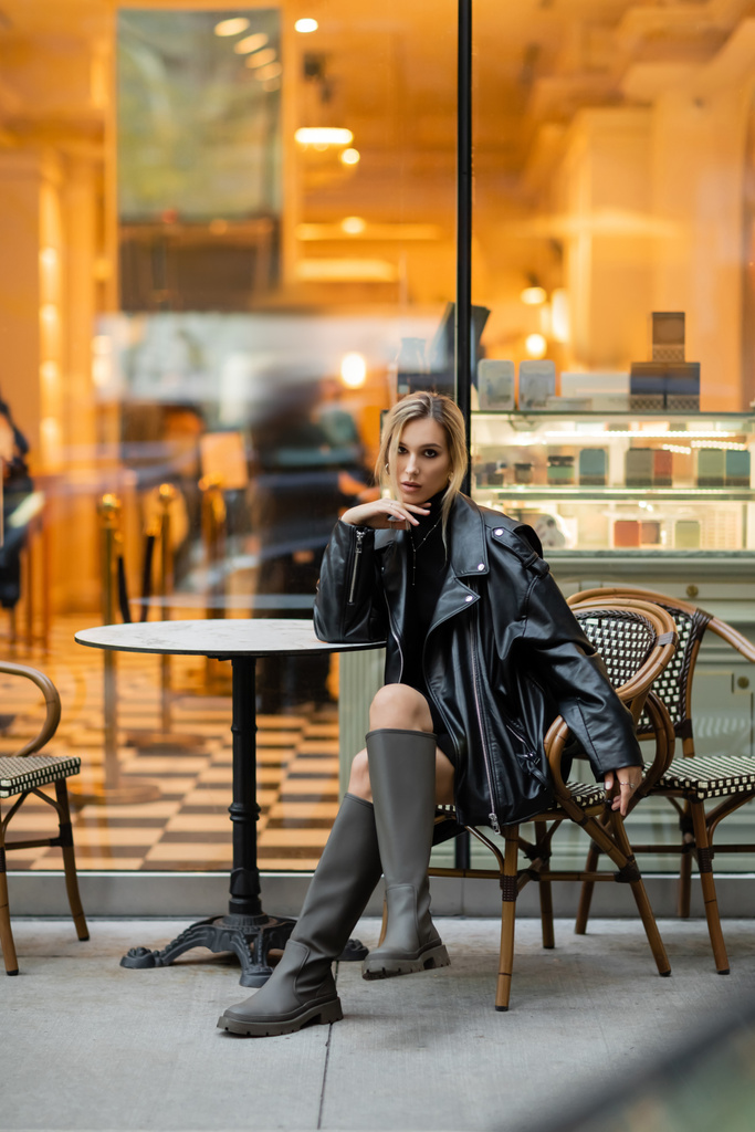 pełna długość blondynki w skórzanej kurtce siedzi w pobliżu okrągłego stołu bistro w kawiarni na świeżym powietrzu w Nowym Jorku  - Zdjęcie, obraz