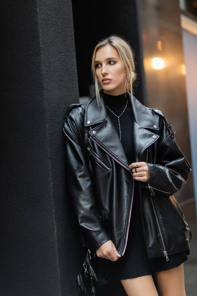 Blondine in stylischer Lederjacke steht mit Handtasche auf der Straße und schaut weg  - Foto, Bild