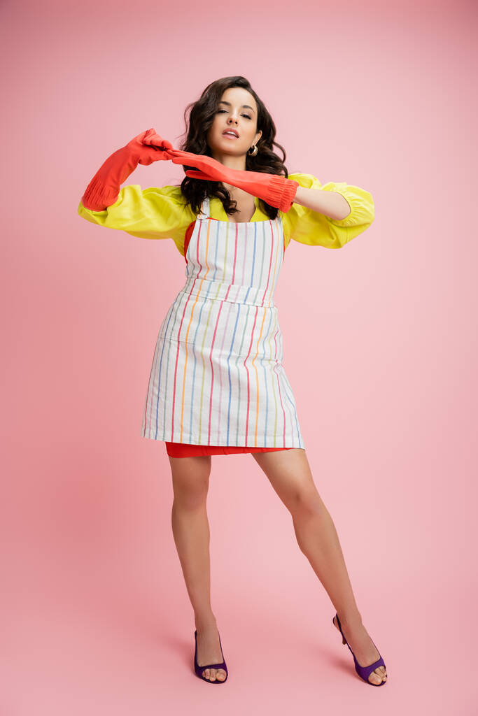 full length of stylish housewife in striped apron taking off red rubber glove while looking at camera on pink background - Photo, Image