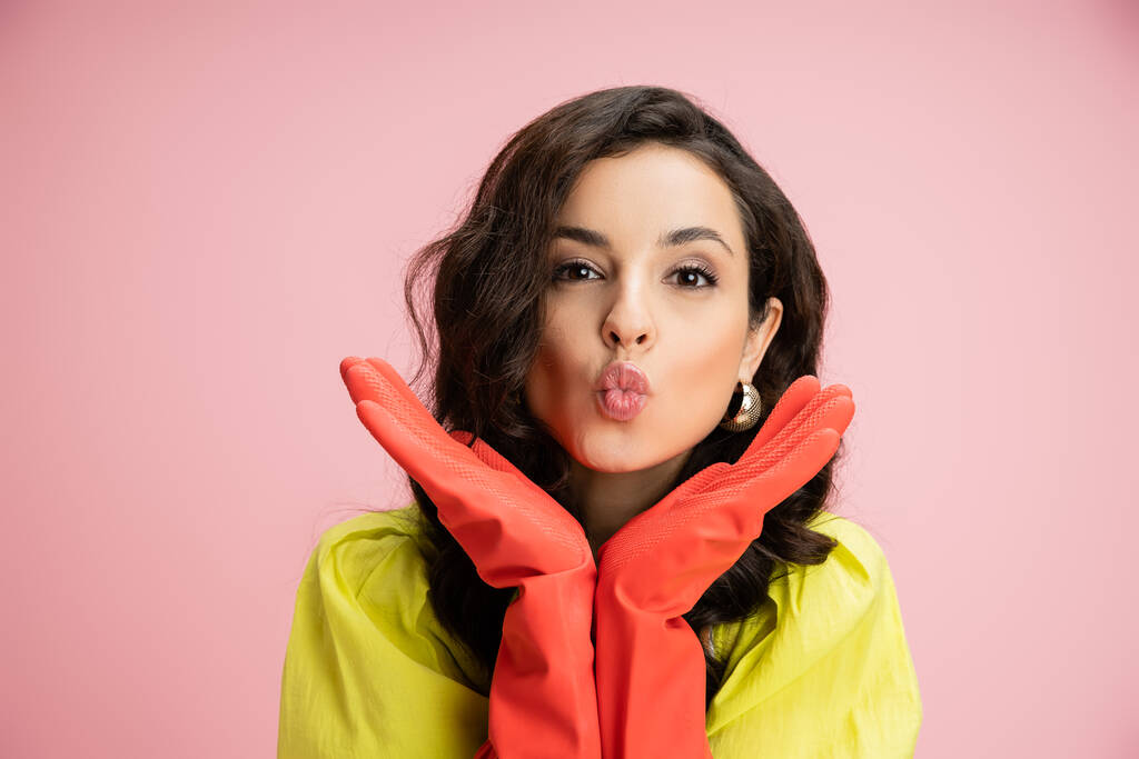 portrait of young brunette housewife in red rubber gloves pouting lips while holding hands near face isolated on pink - Photo, Image