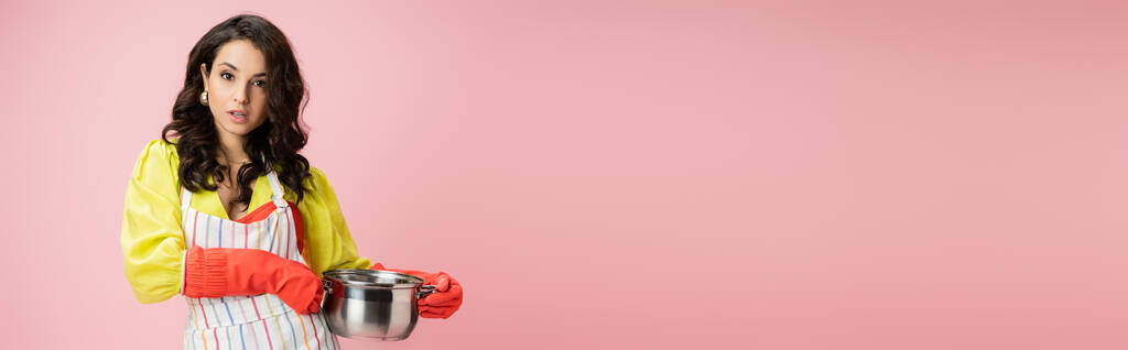 young and pretty housewife holding saucepan and looking at camera isolated on pink, banner - Photo, Image