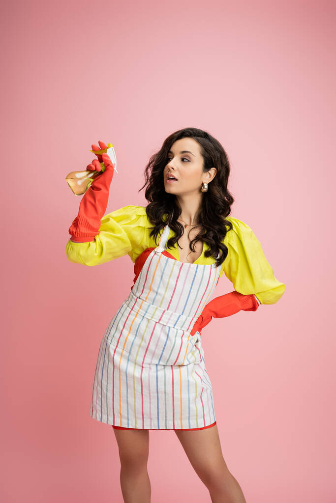 брюнетка домохозяйка в желтой блузке и полосатый фартук держа спрей бутылку и глядя в сторону изолированы на розовый - Фото, изображение