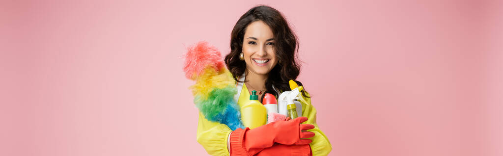 cheerful brunette housewife with colorful feather duster and detergents looking at camera isolated on pink, banner - Photo, Image