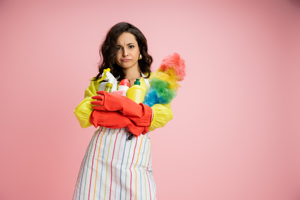 frowning housewife in striped apron and red rubber gloves holding pile of cleaning supplies isolated on pink - Photo, Image