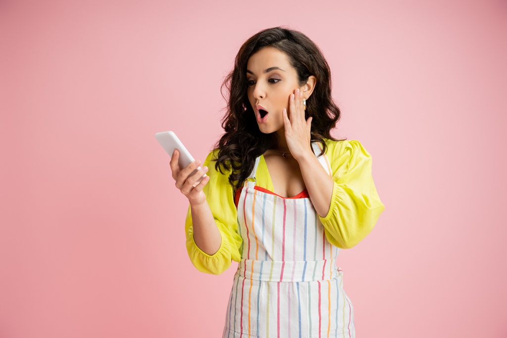 шокированный домохозяйка в желтой блузке и полосатый фартук касаясь щеки, глядя на сотовый телефон изолирован на розовый - Фото, изображение