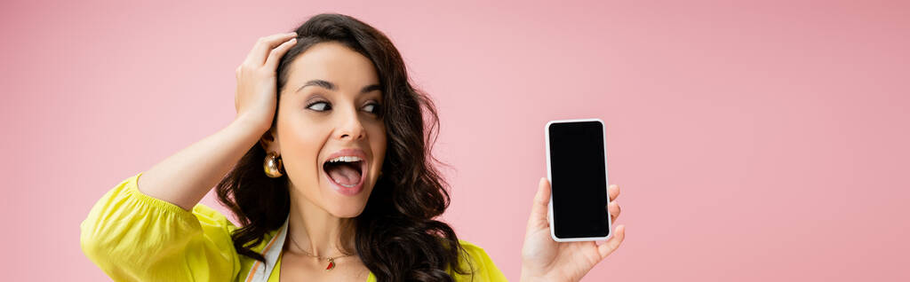 sorprendida ama de casa tocando el pelo ondulado mientras mira el teléfono celular con pantalla en blanco aislado en rosa, pancarta - Foto, Imagen
