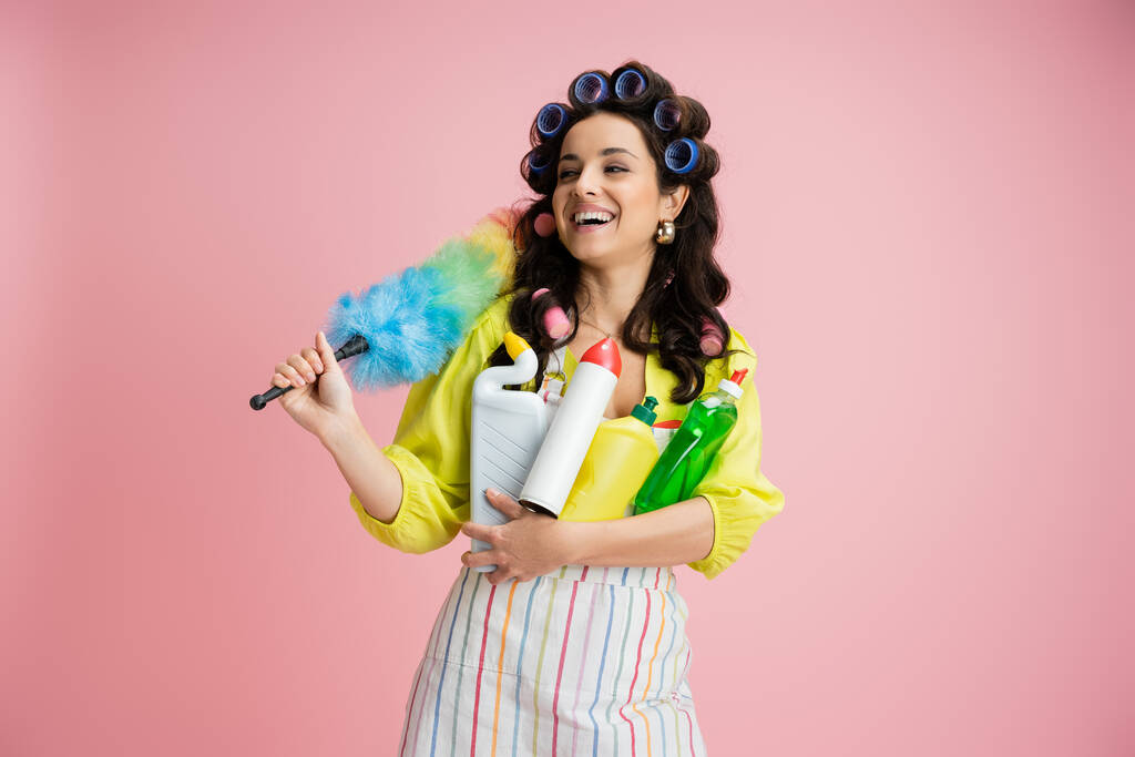 dona de casa alegre em encrespadores de cabelo segurando espanador de penas colorido e suprimentos de limpeza enquanto olha para longe isolado em rosa - Foto, Imagem
