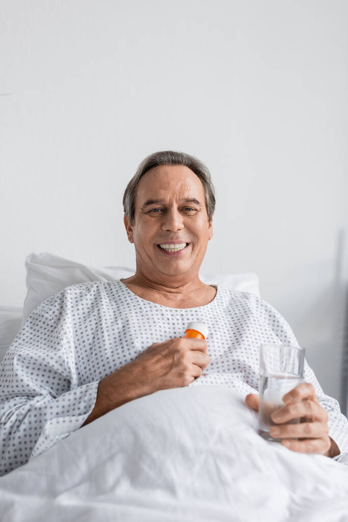 Wesoły siwowłosy mężczyzna trzymający tabletki i wodę patrząc na kamerę na szpitalnym łóżku - Zdjęcie, obraz