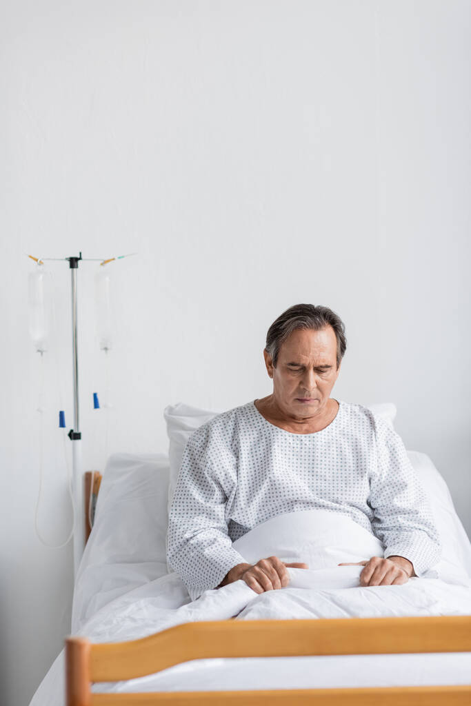 Upset elderly patient in gown sitting on bed near intravenous therapy in clinic - Photo, Image