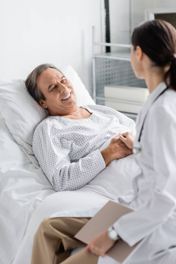 Размытый доктор держит за руку улыбающегося пожилого пациента в клинике - Фото, изображение