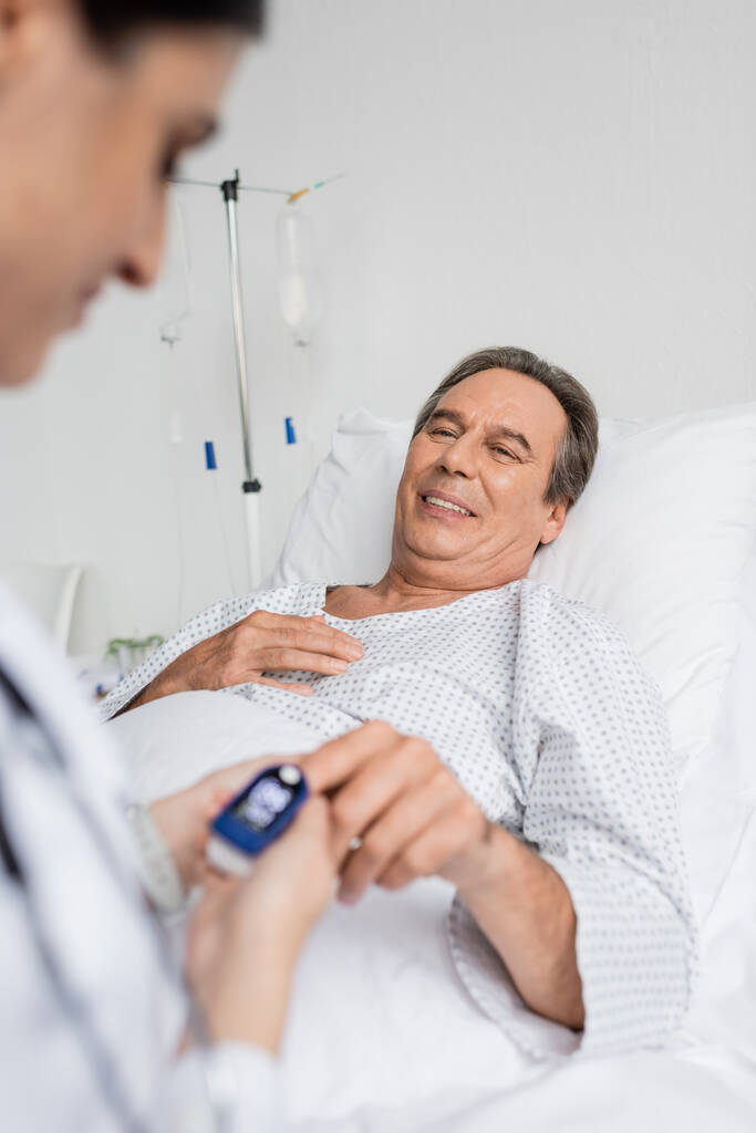 Lachende senior patiënt met polsoximeter op zoek naar wazig arts in kliniek  - Foto, afbeelding