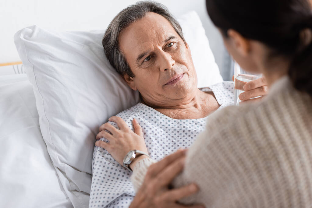 Senior patient holding glass of water and looking at blurred daughter in hospital ward  - Foto, imagen
