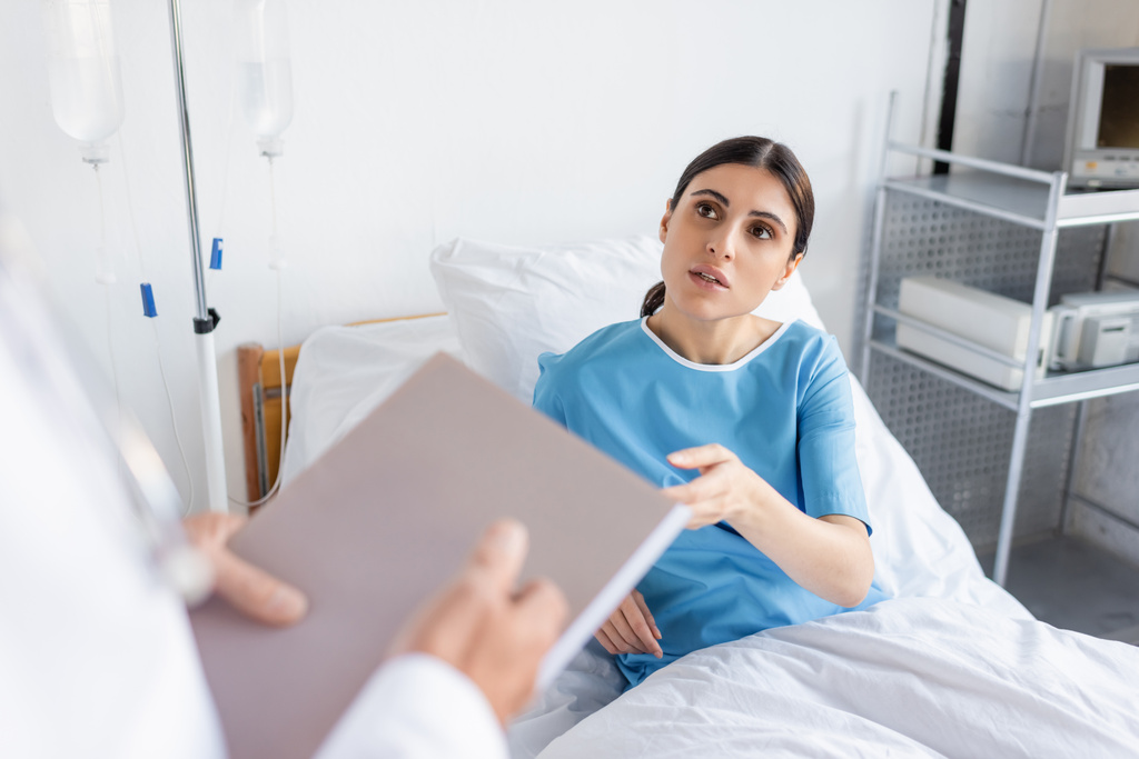 brunette patient talking to blurred doctor with paper folder in hospital ward  - Photo, Image