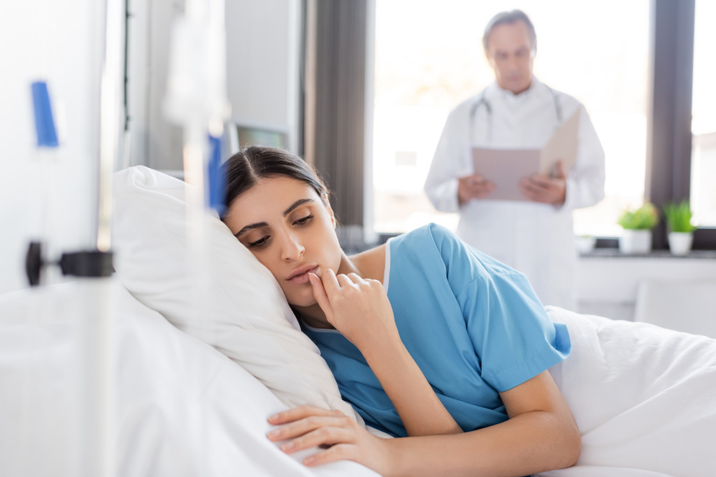 brunette vrouw in patiënt toga liggend op bed in de buurt wazig arts in kliniek  - Foto, afbeelding