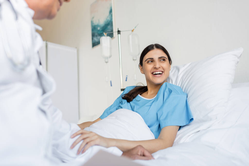 Cheerful patient in gown lying on bed near blurred doctor in clinic  - Photo, Image