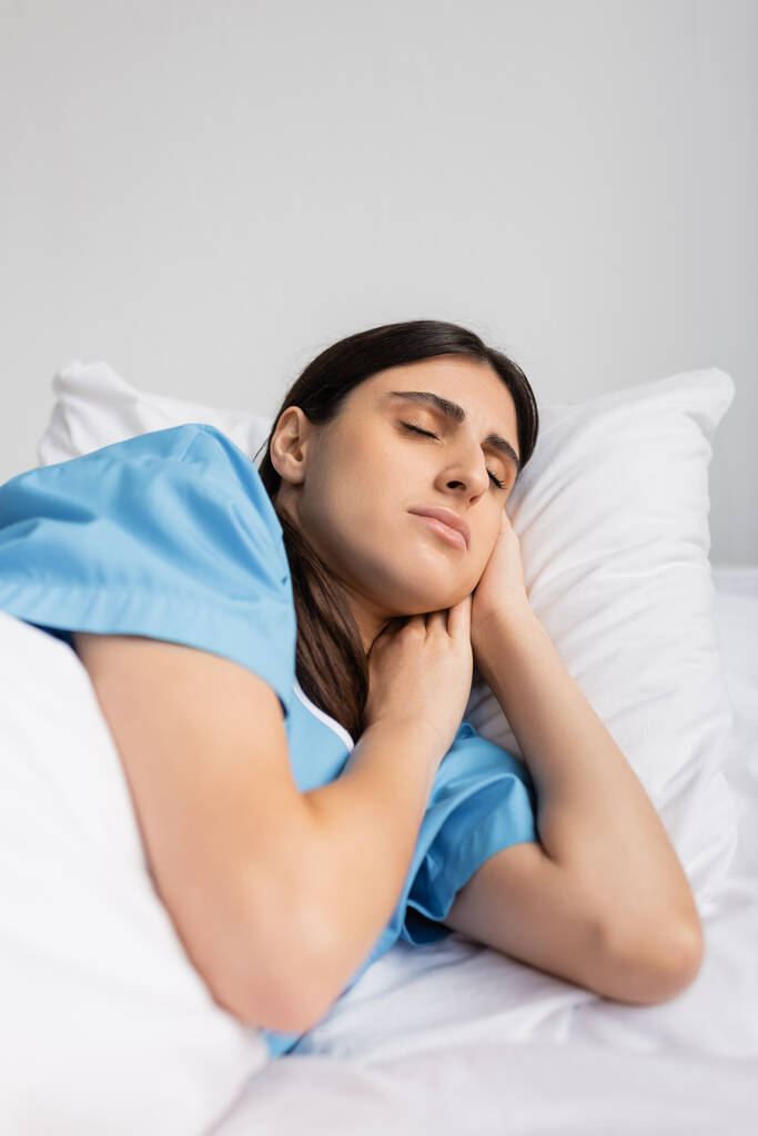 Kranke brünette Patientin im Kleid schläft, während sie in Klinik auf Bett liegt  - Foto, Bild