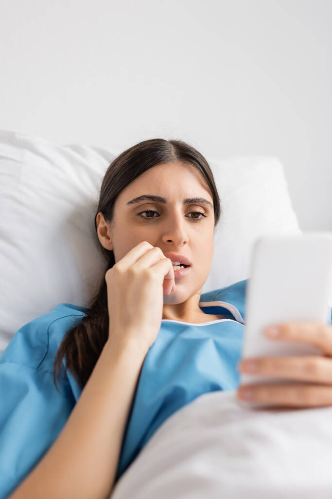 Стрессовый пациент с помощью смартфона, лежащий на кровати в палате больницы  - Фото, изображение