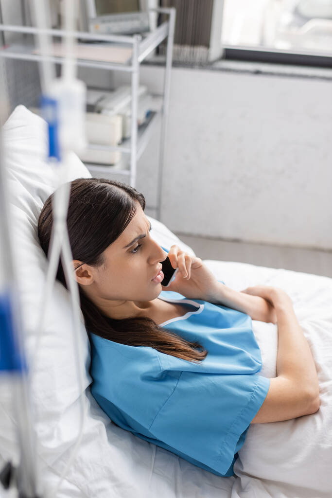 pacjent rozmawiający na smartfonie podczas leżenia na łóżku w pobliżu nieostrej terapii dożylnej w szpitalu  - Zdjęcie, obraz
