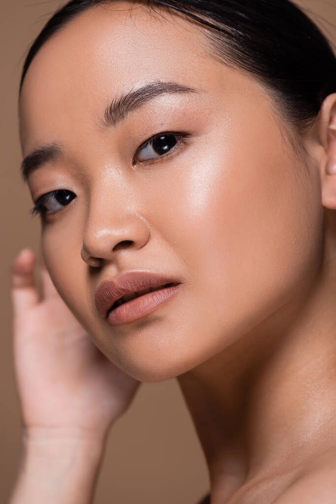 Портрет молодой азиатки с натуральным макияжем, смотрящей в камеру изолированную на коричневом  - Фото, изображение