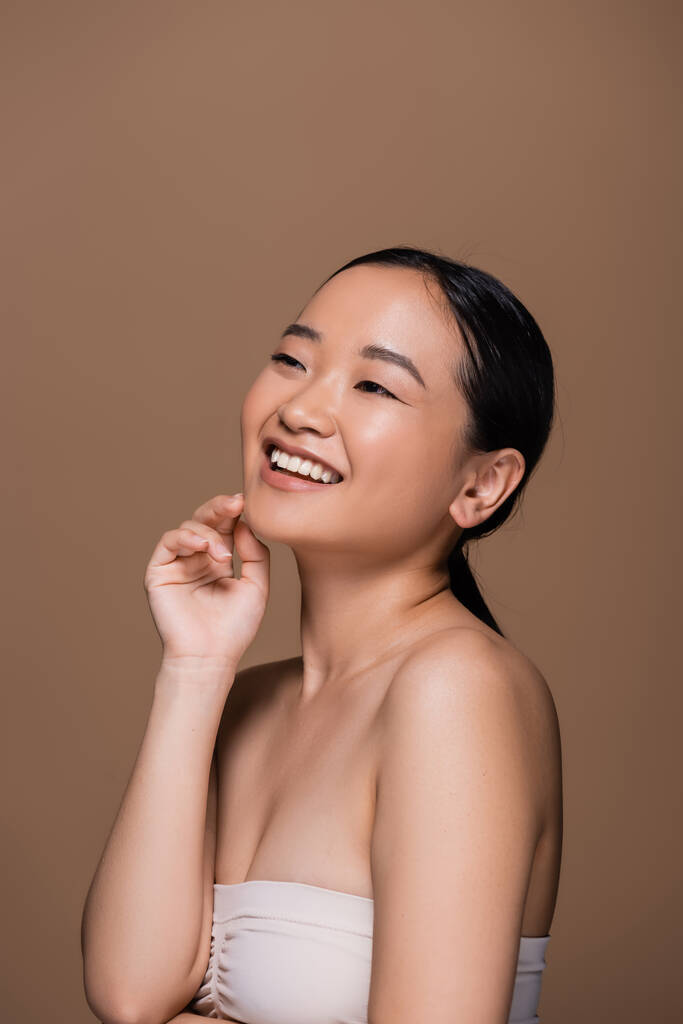 Positive asian woman in top touching face and looking away isolated on brown  - Photo, image