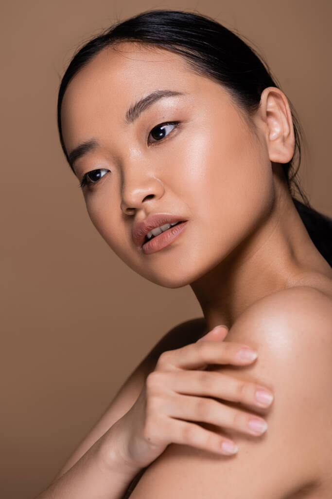 Молодая азиатская женщина с натуральным макияжем касаясь обнаженного плеча изолированы на коричневый  - Фото, изображение