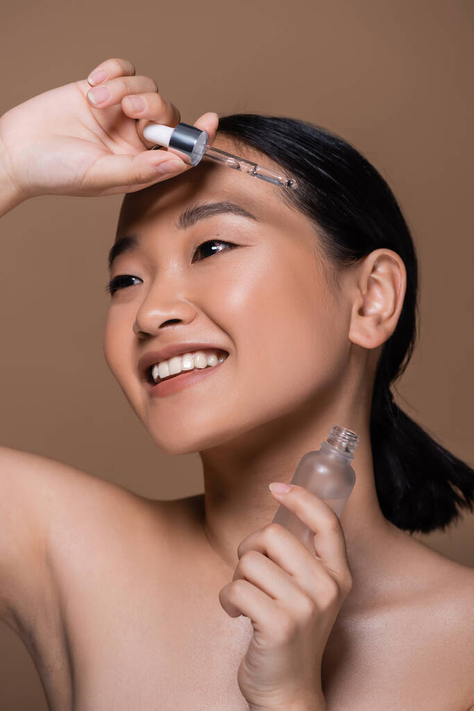 Smiling asian woman with naked shoulders holding cosmetic serum isolated on brown  - Fotoğraf, Görsel