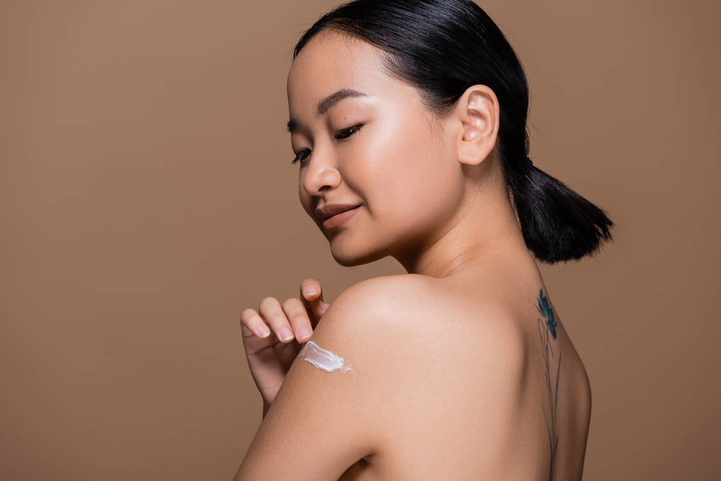 Tattooed asian woman with naked shoulders applying cosmetic cream on shoulder isolated on brown  - Foto, imagen