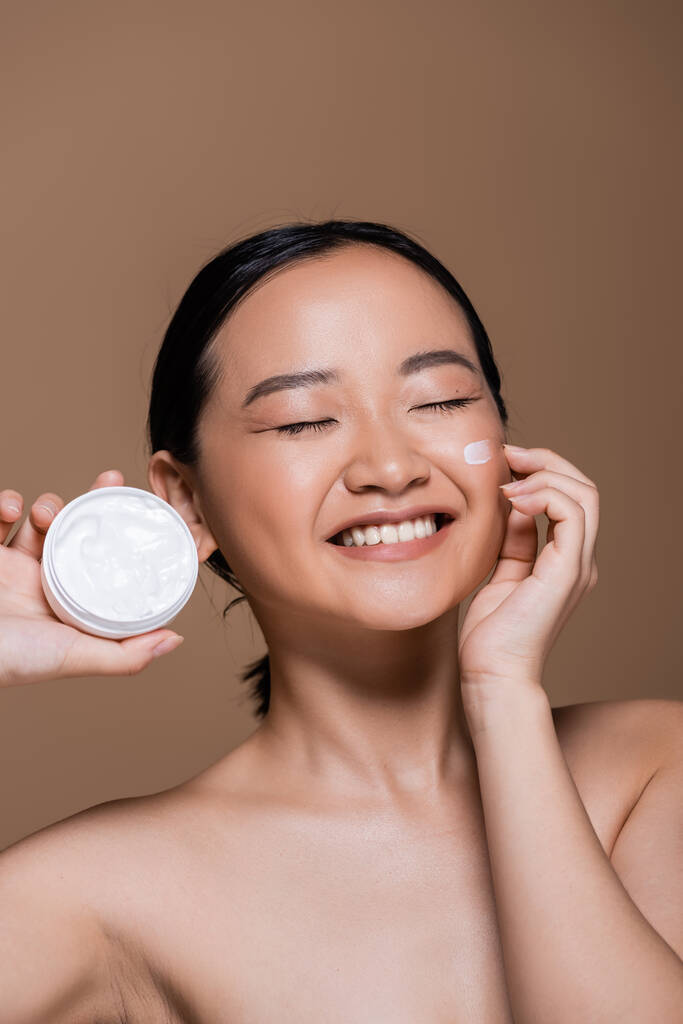 Pretty asian woman with naked shoulders holding cosmetic cream isolated on brown  - Fotoğraf, Görsel