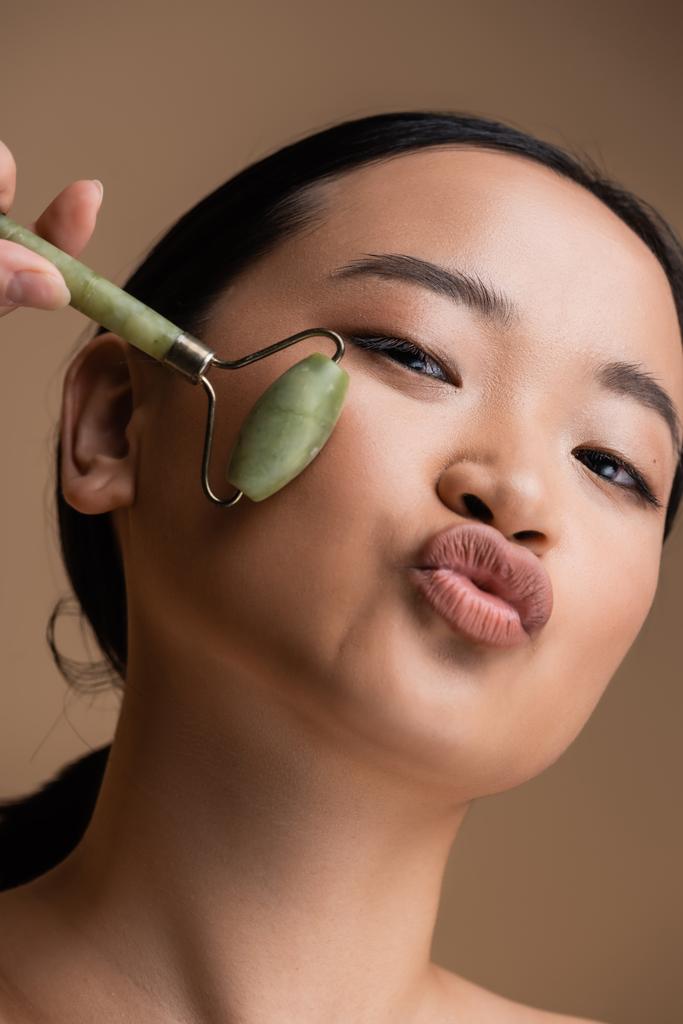 Portrait of asian woman holding jade roller and pouting lips isolated on brown  - Foto, immagini