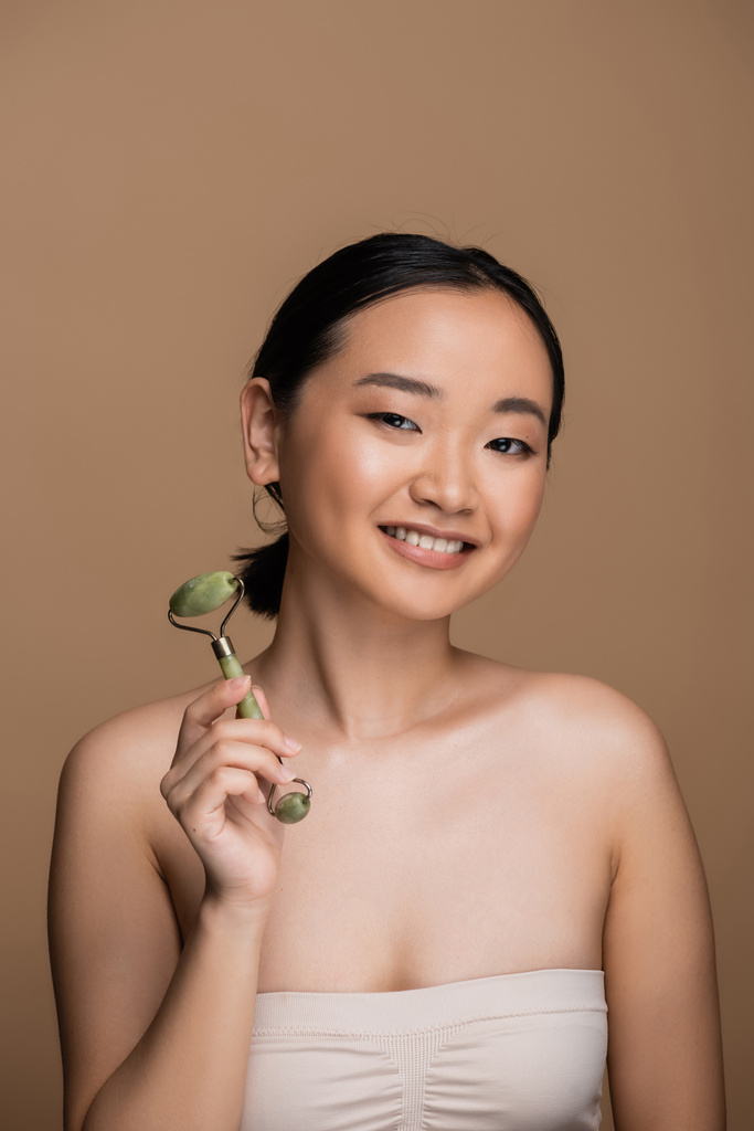 Positive asian model in beige top holding jade roller and looking at camera isolated on brown  - Valokuva, kuva