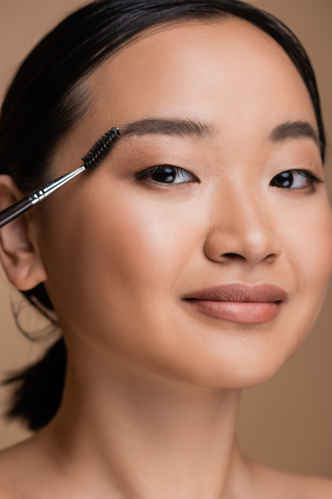 Портрет молодой азиатки с макияжем, держащей кисть бровей изолированной на коричневом  - Фото, изображение
