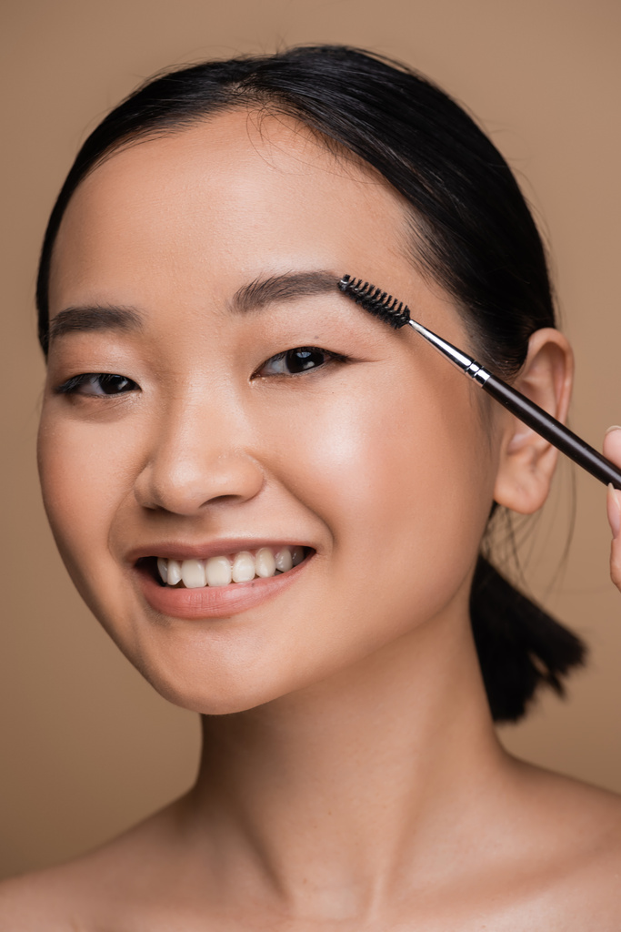 Pretty asian model styling eyebrow while holding brush isolated on brown  - Foto, imagen