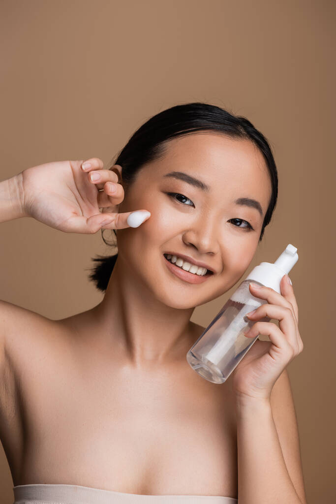 Positive asian model with naked shoulders holding cleanser with dispenser isolated on brown  - Fotografie, Obrázek