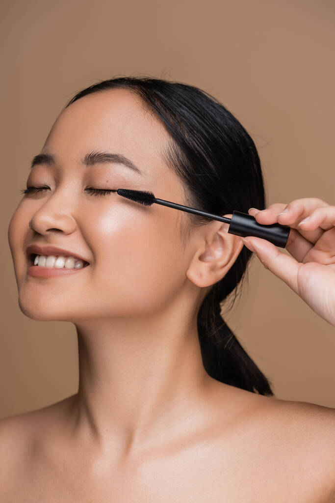 Cheerful asian woman with naked shoulders holding mascara applicator isolated on brown  - Zdjęcie, obraz