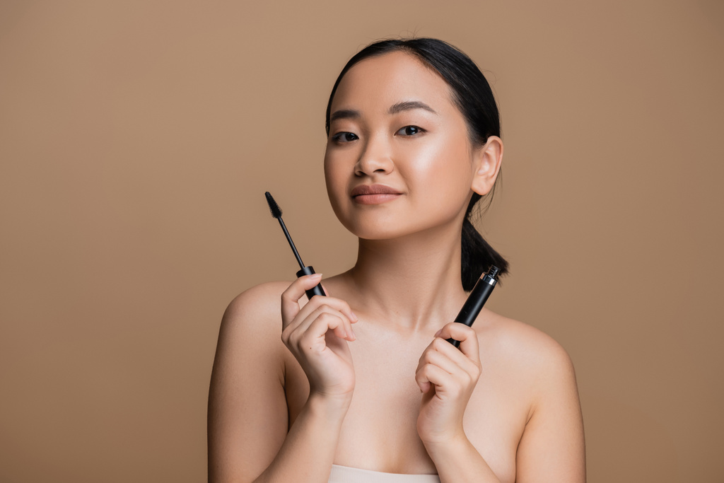 Pretty asian woman holding mascara and looking at camera isolated on brown  - Foto, imagen