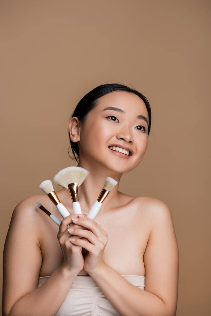 Positive asian model in top holding cosmetic brushes isolated on brown  - Foto, immagini