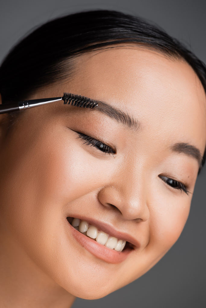 自然メイクブラッシング眉毛を持つ陽気なアジアの女性の肖像画は灰色に隔離された - 写真・画像