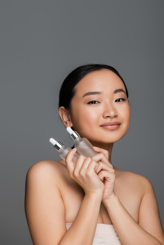 брюнетка азиатская женщина с идеальной кожей держа бутылки натуральной косметической сыворотки изолированы на сером - Фото, изображение