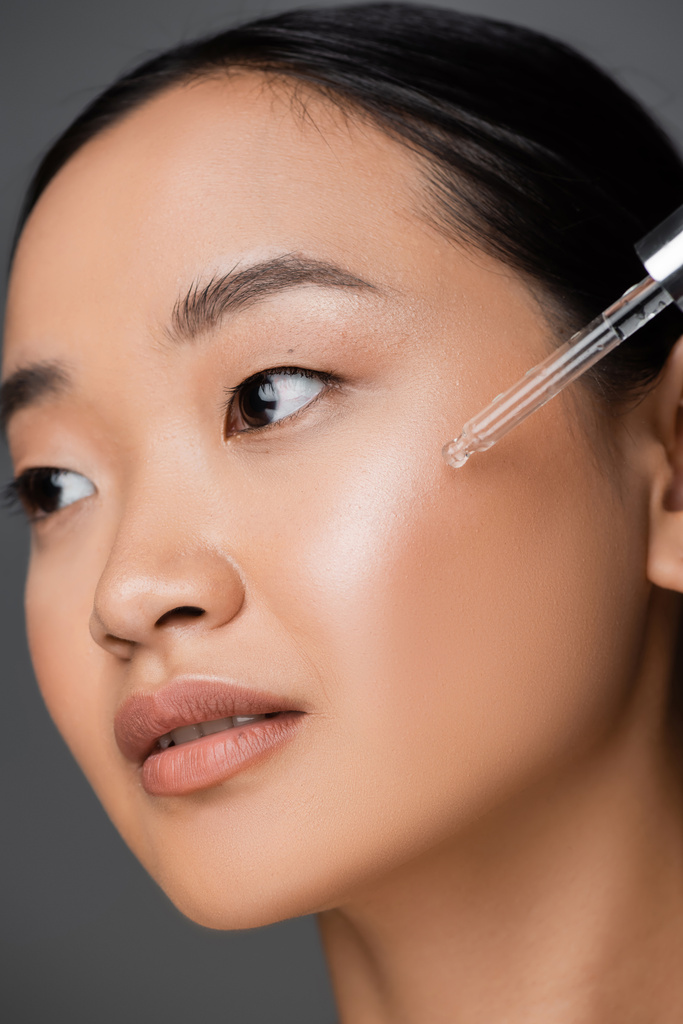 close up view of asian woman applying moisturizing cosmetic serum during facial pampering isolated on grey - Fotografie, Obrázek