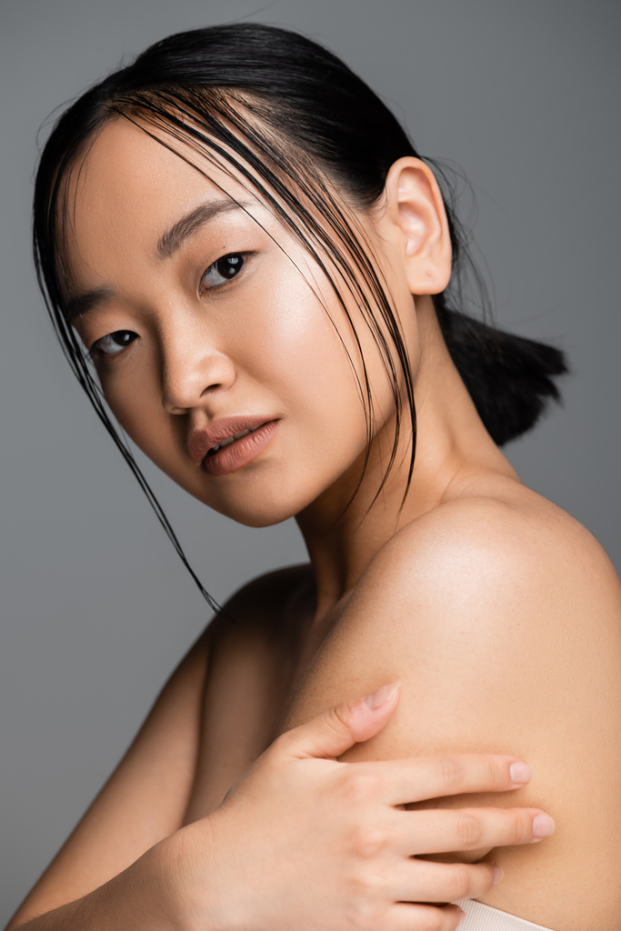 portrait of brunette asian woman with natural makeup touching bare shoulder and looking at camera isolated on grey - Photo, Image