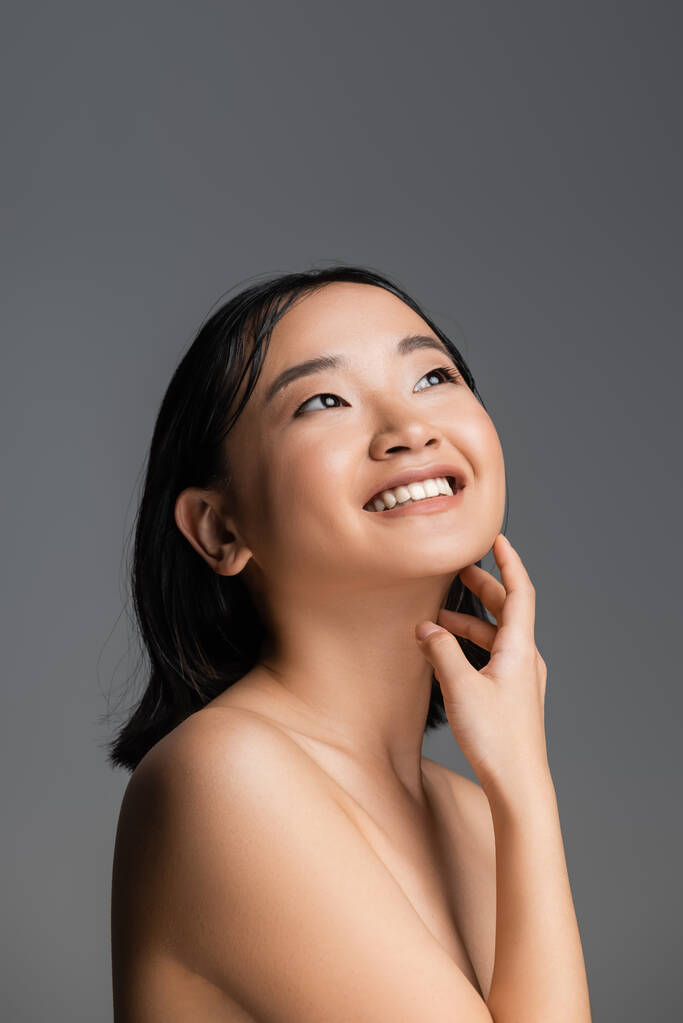 cheerful asian woman touching perfect skin and looking away isolated on grey - Foto, Imagem