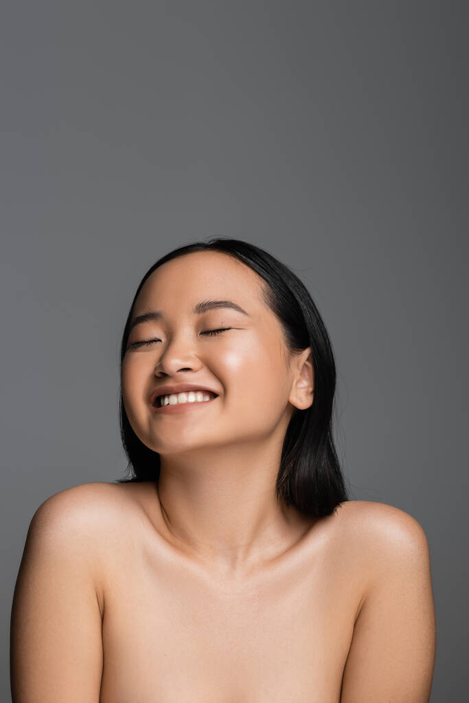 cheerful asian woman with bare shoulders and natural makeup smiling with closed eyes isolated on grey - Foto, imagen