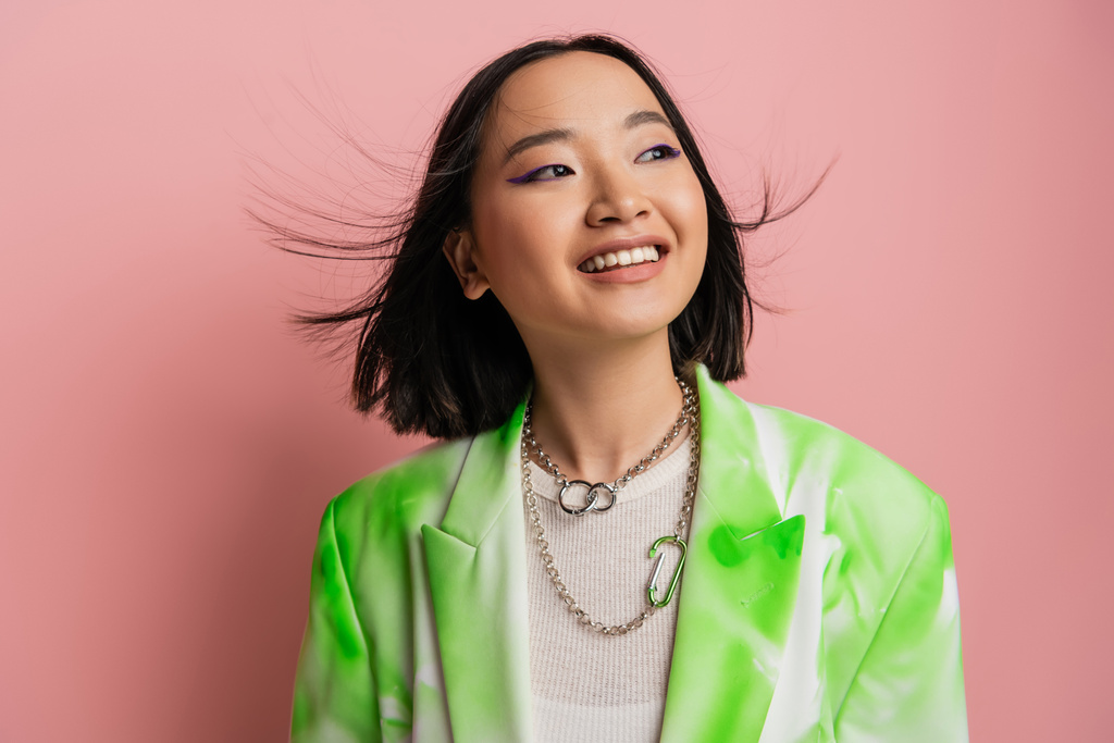 joyful asian woman in trendy outfit and makeup with blue eyeliner looking away on pink background - Foto, imagen
