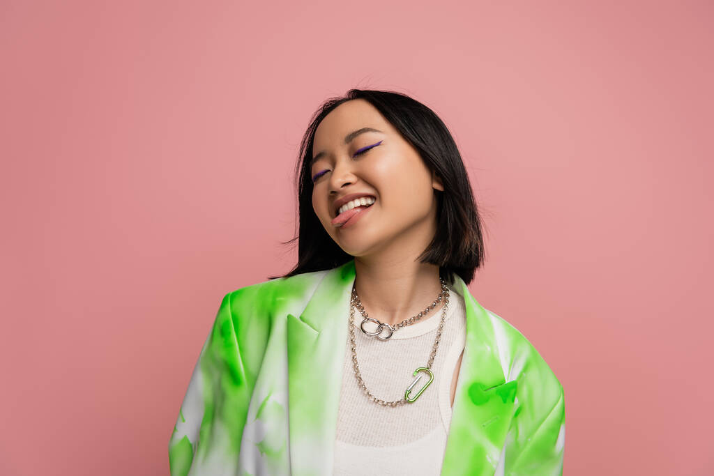 cheerful asian woman in green and white jacket and metal necklaces sticking out tongue isolated on pink - Foto, Imagen