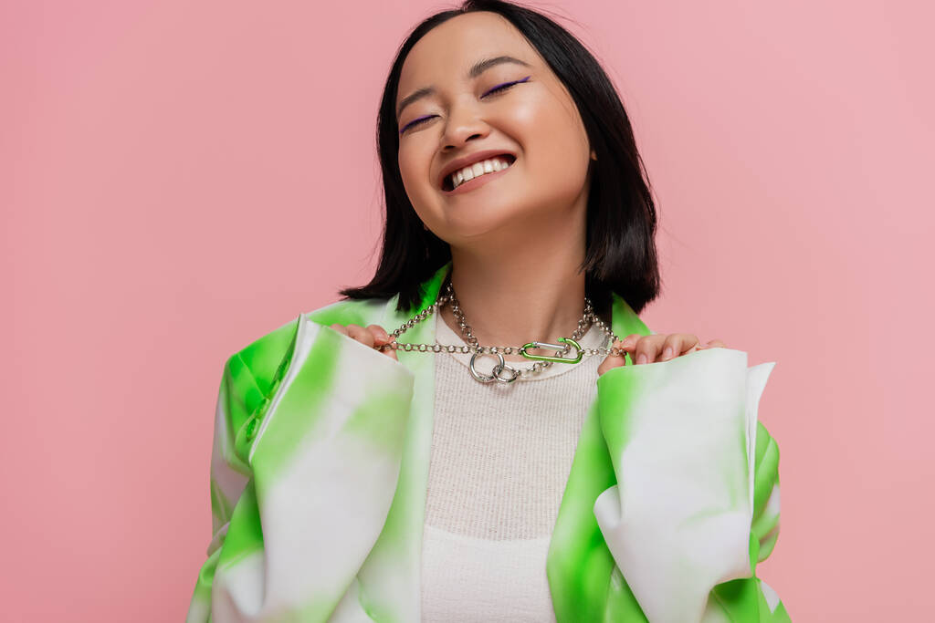 pleased asian woman in green and white clothes touching silver necklaces isolated on pink - Foto, Imagem