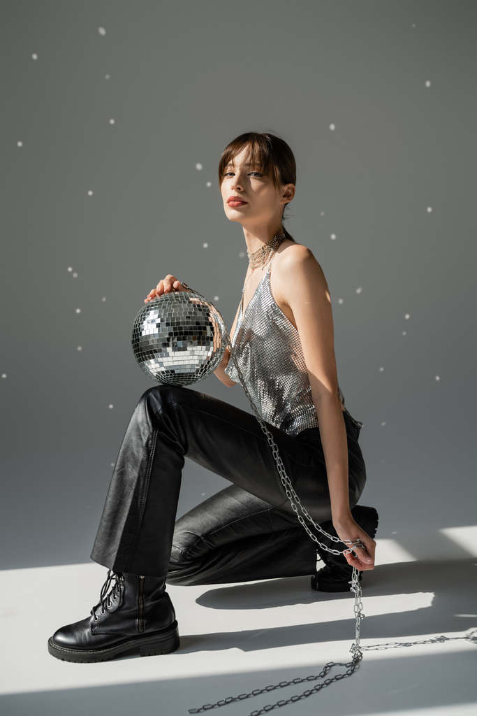 full length of young woman in trendy top and leather trousers holding chain with shiny disco ball on grey  - Foto, immagini